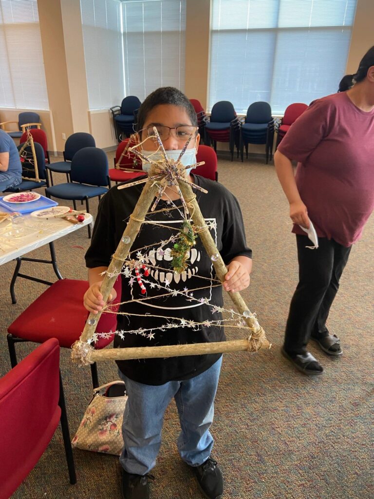 An artist holds a completed stick tree decorated with a straw star at the top, a silver star garland, a beaded candy cane and a cluster of artificial pine at the centre.