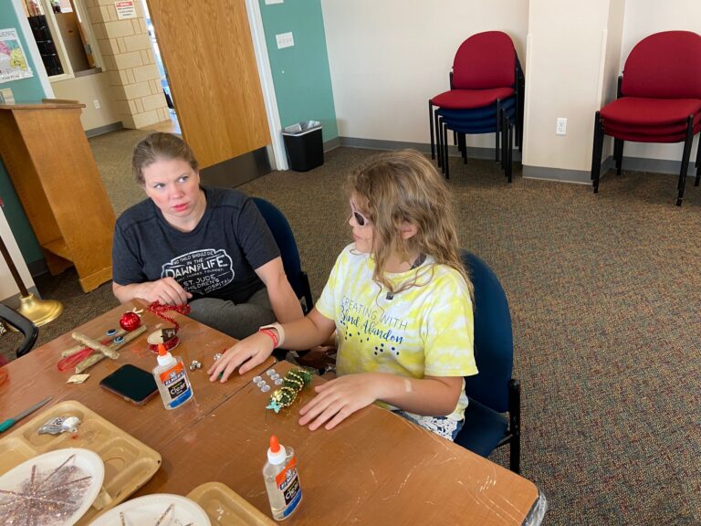 An artist and a volunteer work side by side at a table, sorting red ornaments, glitter picks and glue. Wooden trays of branch pieces and craft supplies cover the tabletop.