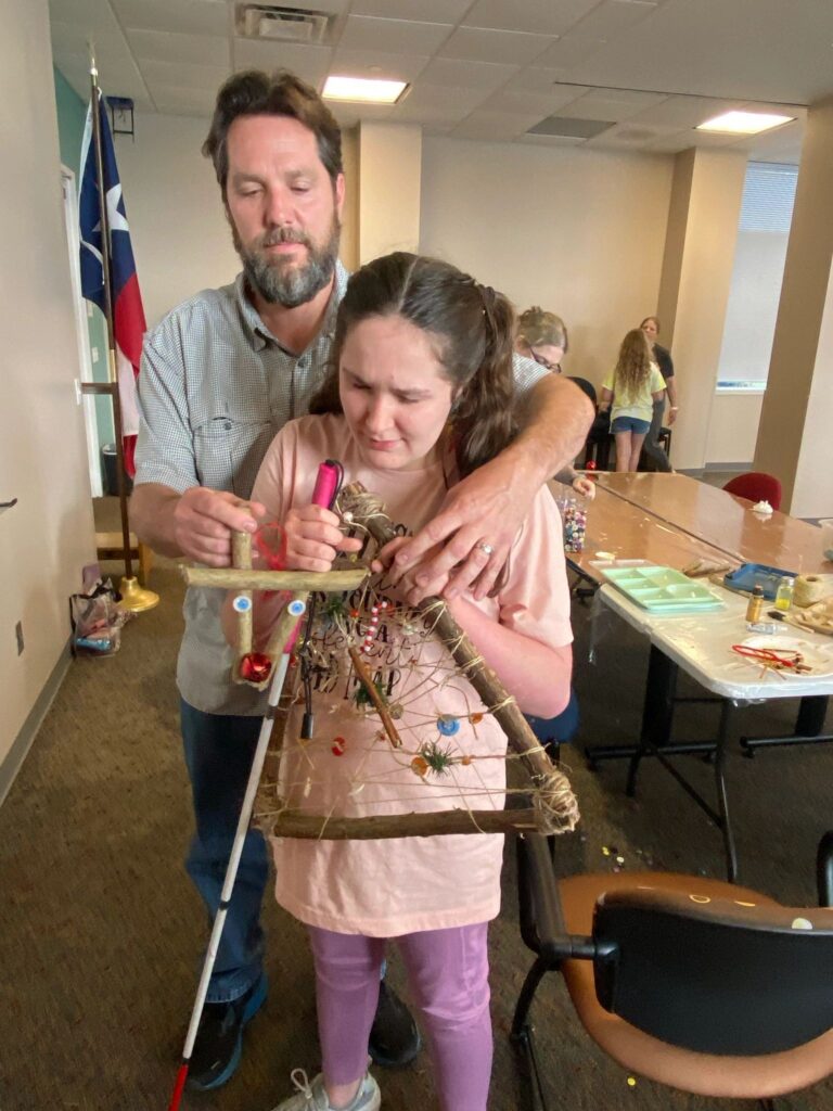 A volunteer stands behind an artist, guiding their hands across a finished stick tree so they can feel the twine rows, beads and greenery. The artist's white cane rests beside them.