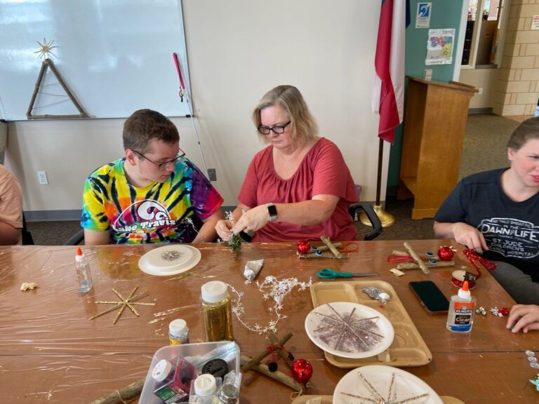 Artists and a volunteer at a table gluing sprigs of greenery and glitter stars onto their pieces. Paper plates hold gold glitter starbursts drying in front of them.