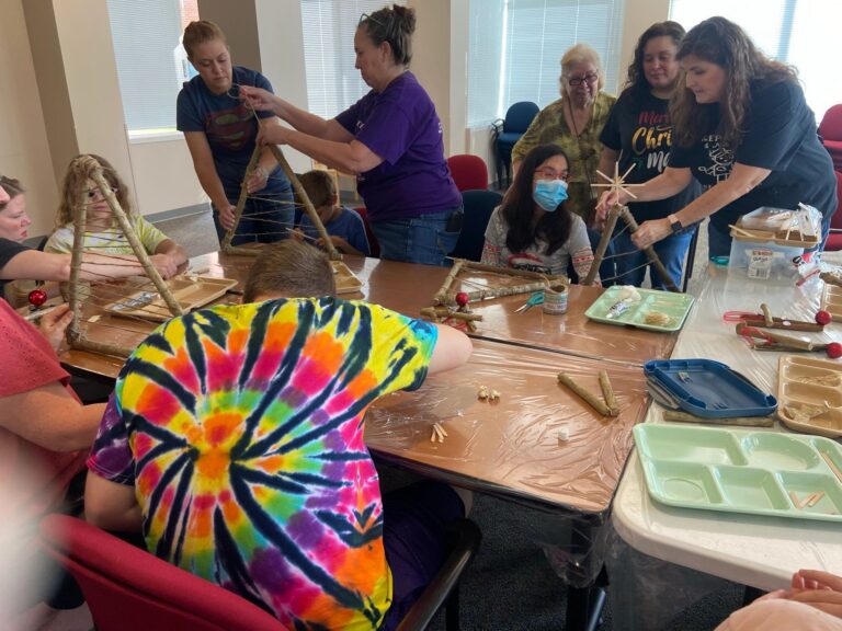 Artists and volunteers gathered around a table, several holding triangular branch frames upright as twine is wound between the sides. Trays of cinnamon sticks and craft materials are spread across the table.