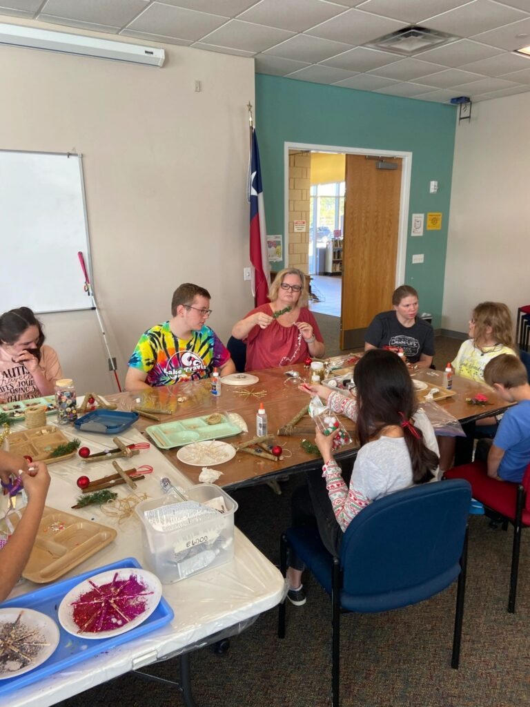 A wide view of the session, with artists working around a long table on their stick trees. Trays of greenery, red ornaments, glue and beads are spread out, and a glitter starburst dries on a plate in the foreground.
