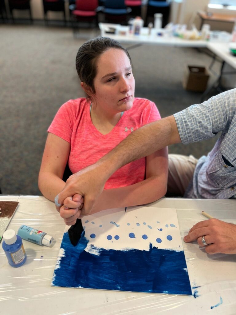 A volunteer helps an artist paint a row of blue dots above a dark blue painted band on a canvas.