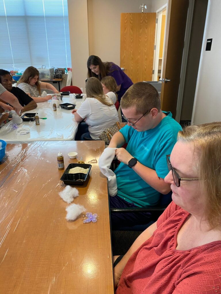Artists and a volunteer at a table gluing charms onto sock bunnies, with a cotton-stuffed lavender charm and craft supplies spread across the table.