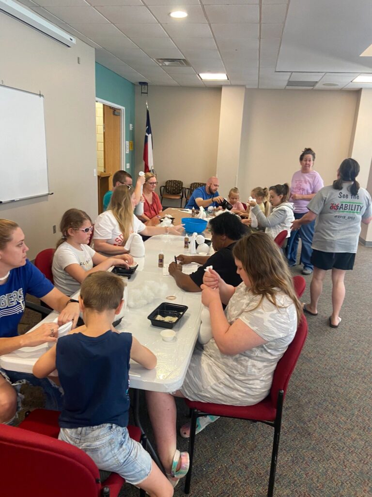 A wide view of the studio, with artists and volunteers gathered around several tables making sock bunnies, rice and cotton spread across the tabletops.