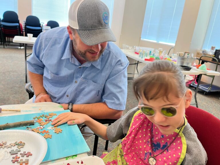 A volunteer helps an artist arrange gingerbread-coloured puzzle pieces onto a canvas with a twig trunk.