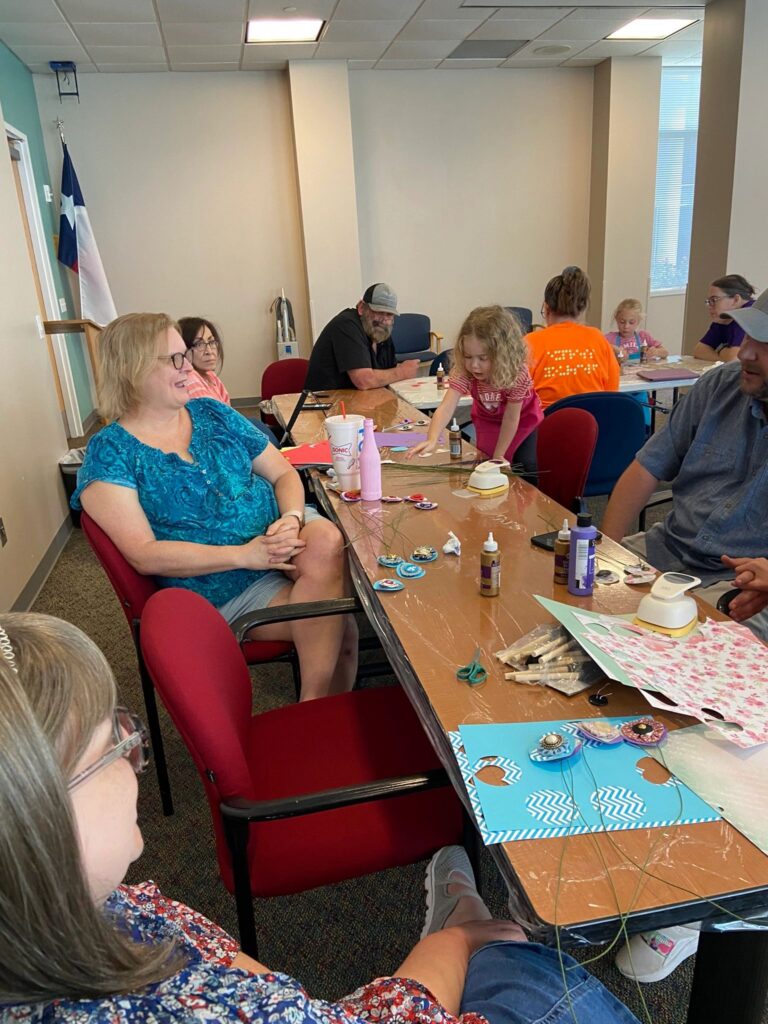 A wide view of the studio, with artists and volunteers seated around several tables working on painted bottles decorated with paper flowers.