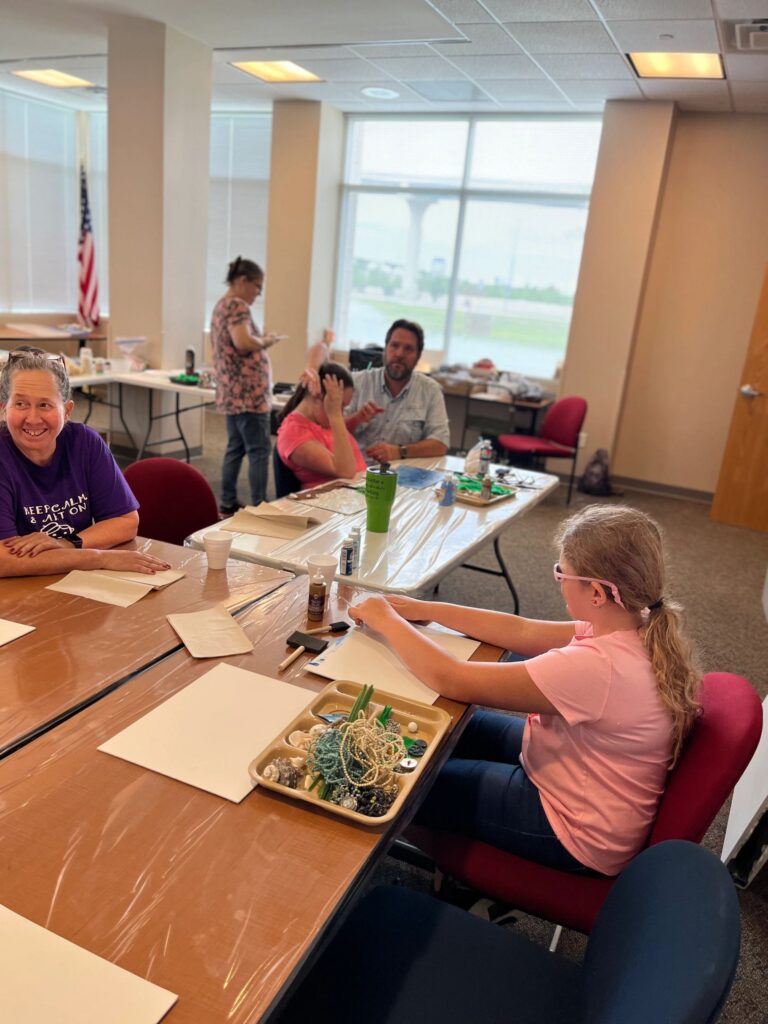A wide view of the studio, with artists and volunteers seated at several tables working on beach-themed canvases.