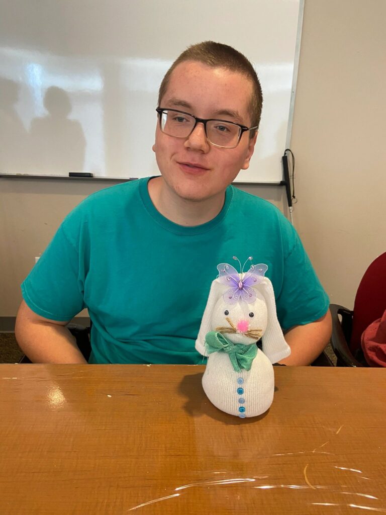 An artist sits at a table with a finished white sock bunny decorated with a purple glitter butterfly and a pink pom-pom nose.