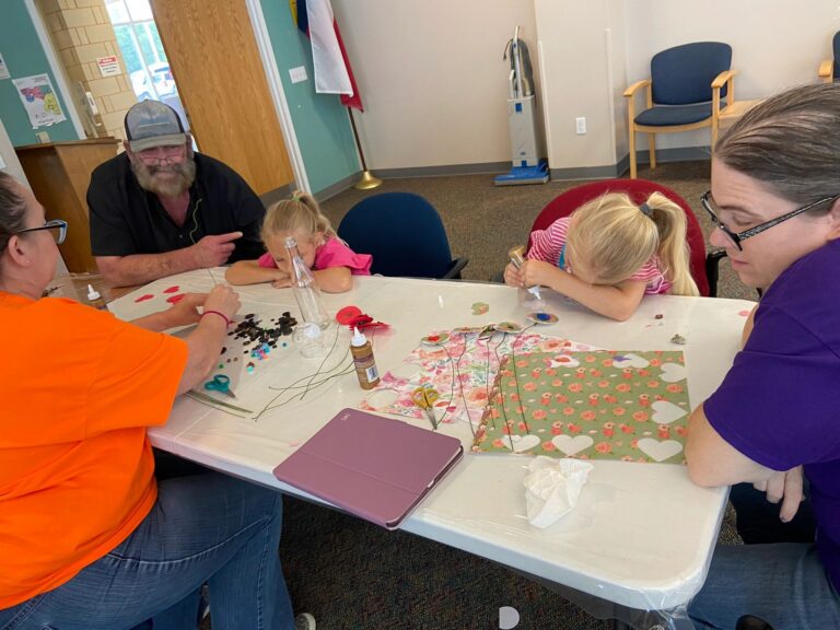 A wide view of the studio, with artists and volunteers seated at tables making sock bunnies from white socks stuffed with rice and cotton.