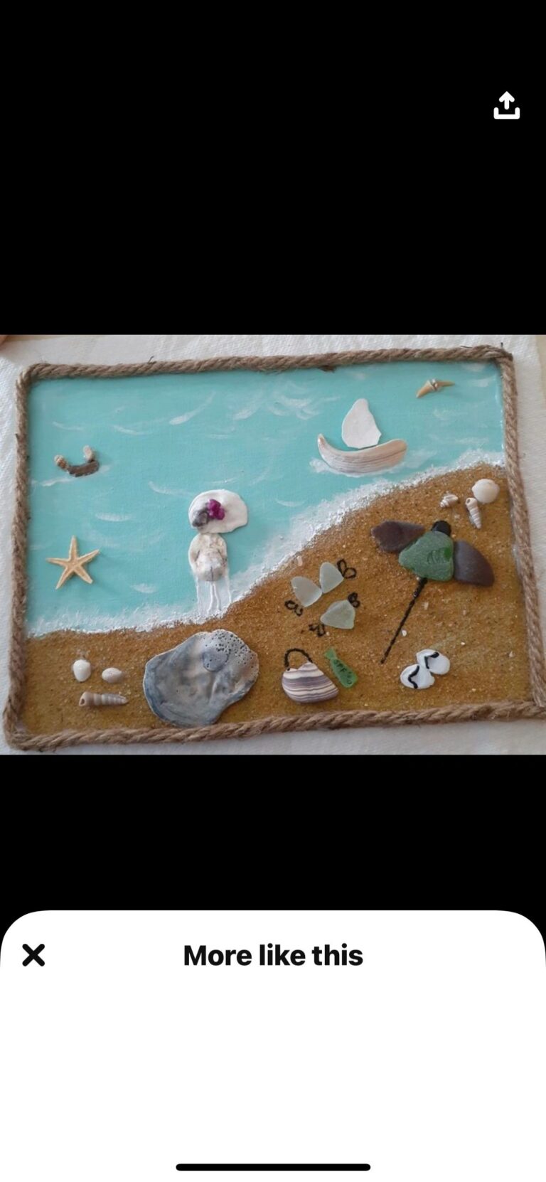 A framed pebble-art beach scene shown on a phone screen, with a shell-and-cloth figure, tiny shell sailboat, sea glass butterflies and a mermaid's purse charm on a sandy shore, used as inspiration for the day's craft.