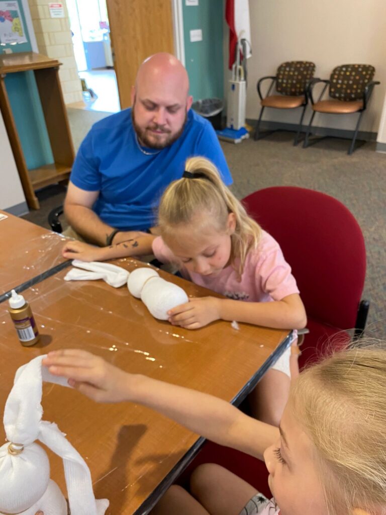 An artist presses their hands into a partly finished sock shape at the table, a volunteer looking on and another sock bunny visible nearby.
