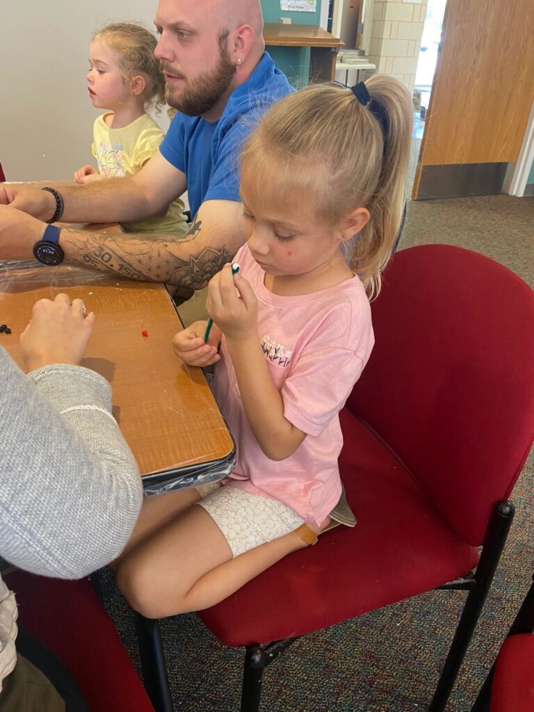A young artist threads a green bead onto a pipe cleaner at a table, seated among other artists and a volunteer.