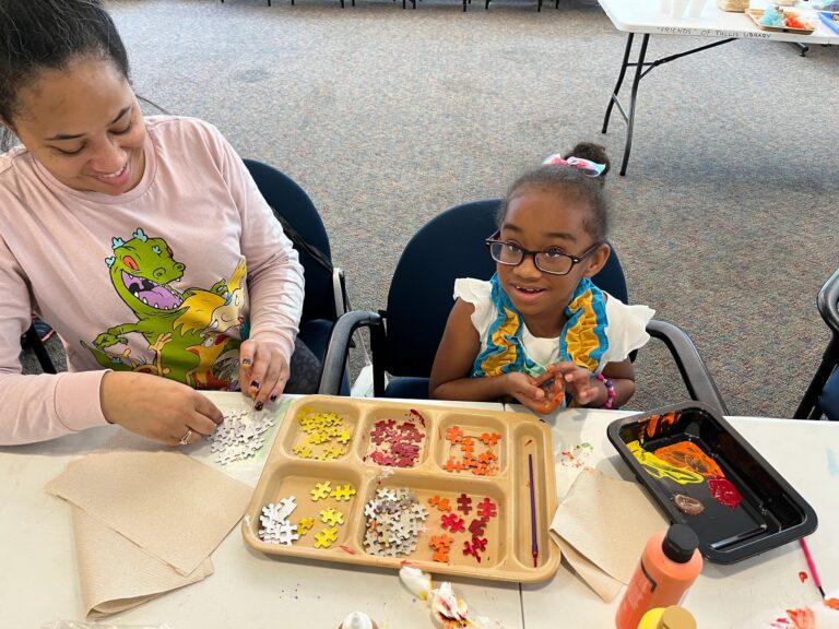 An artist sorts puzzle pieces into a divided tray by colour, with an orange paint tray nearby on the table.