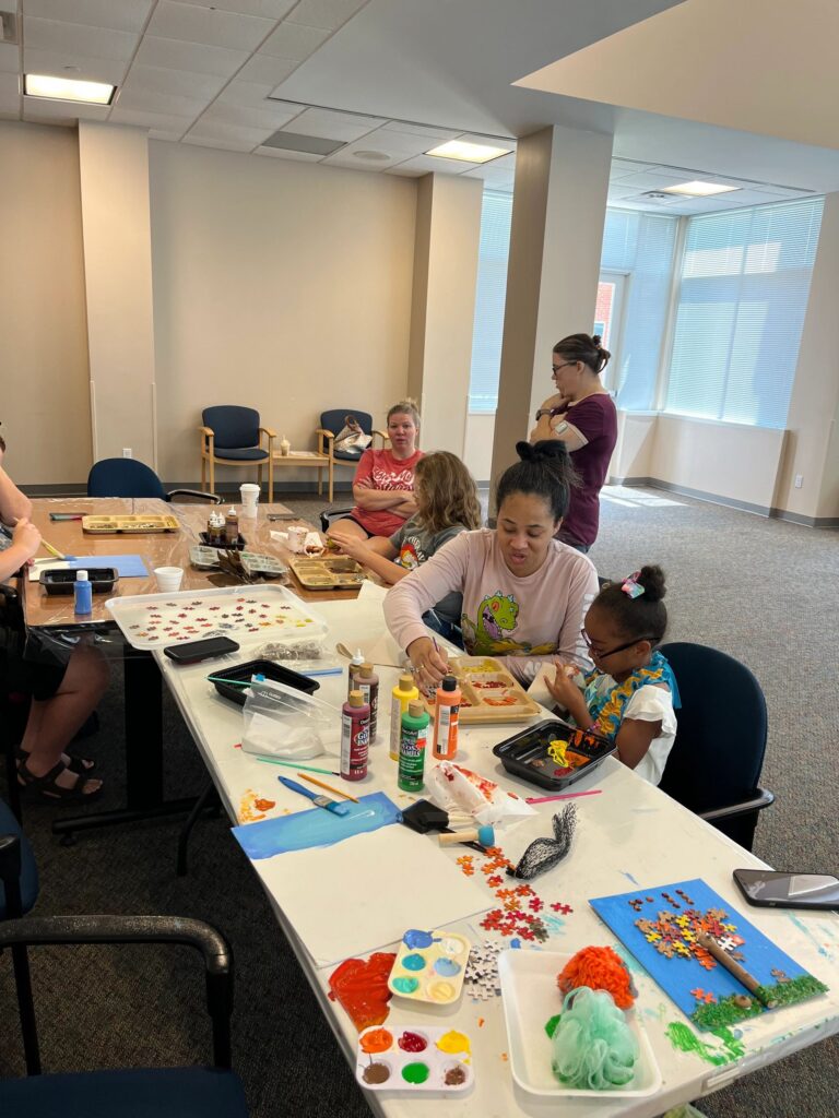 A wide view of the studio, with artists and volunteers seated around a long table covered in paint trays, puzzle pieces and materials for the fall tree canvases.