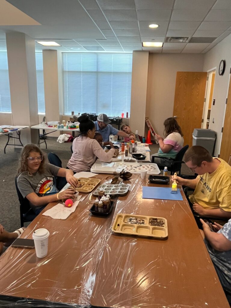 A wide view of the studio, with artists and volunteers gathered around several tables covered in craft supplies for the session.
