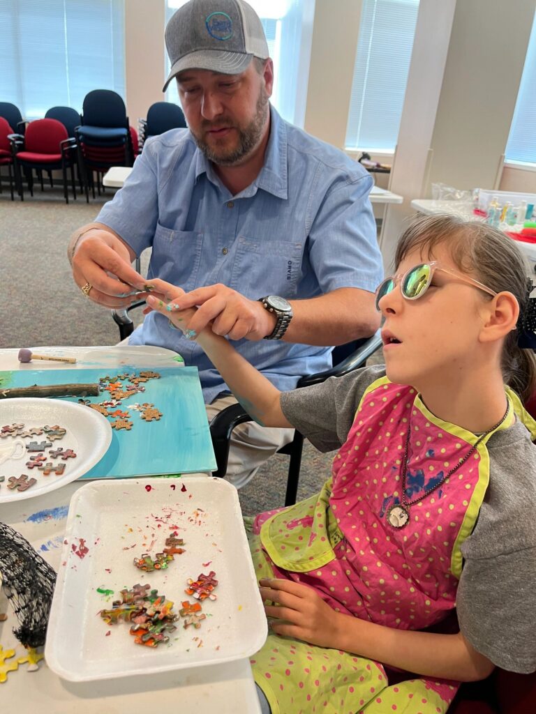 A volunteer helps an artist arrange puzzle pieces and beads on a fall tree canvas, with a plate of loose pieces set on the table.