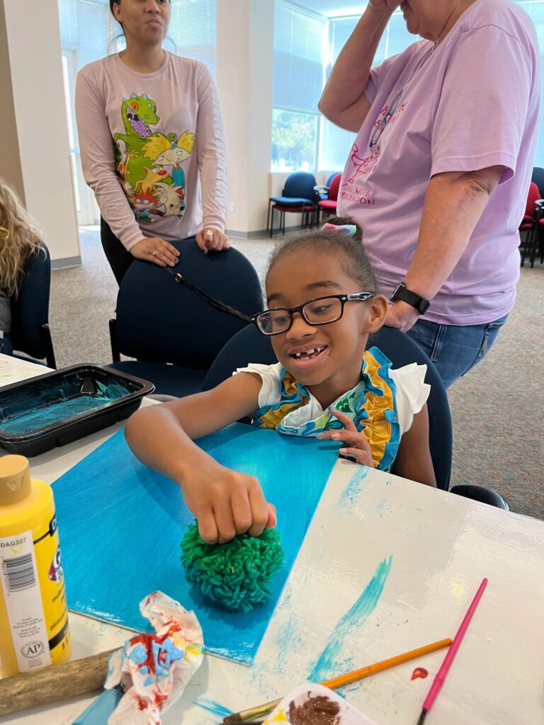 A volunteer helps an artist trace fingertips through green paint on a partly finished blue and white canvas.