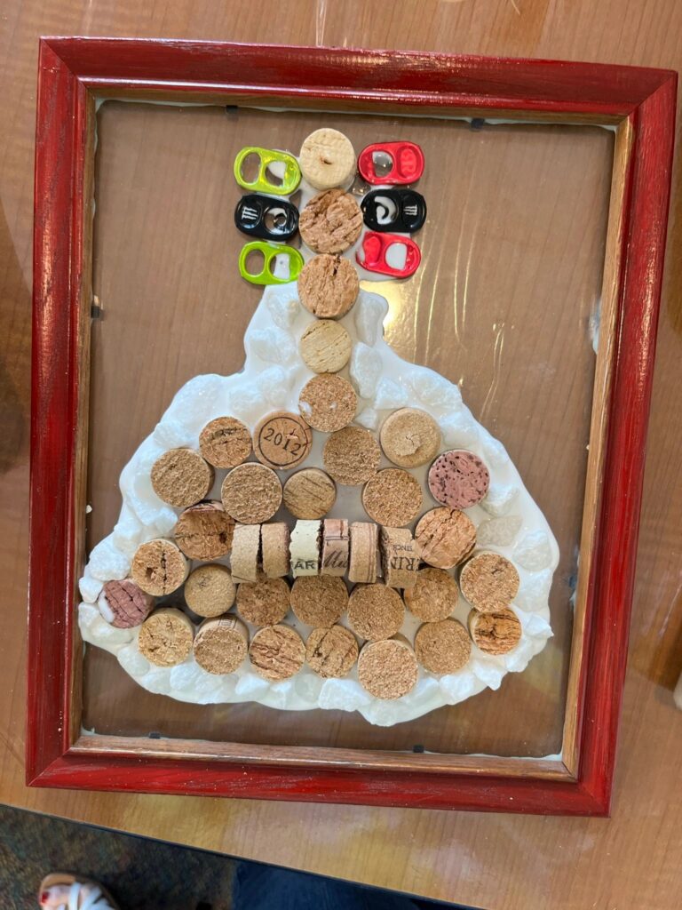 A framed cork art piece shaped like a Christmas tree, with pull-tabs arranged as a star on top and corks stacked below, one marked with the year 2012.