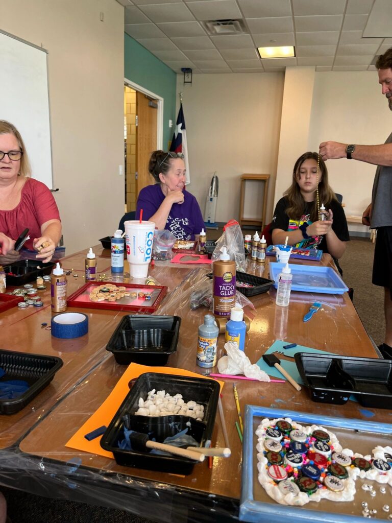 A wide view of the cork art table, with artists and volunteers arranging corks, pull-tabs and beads on framed boards, tacky glue bottles within reach.