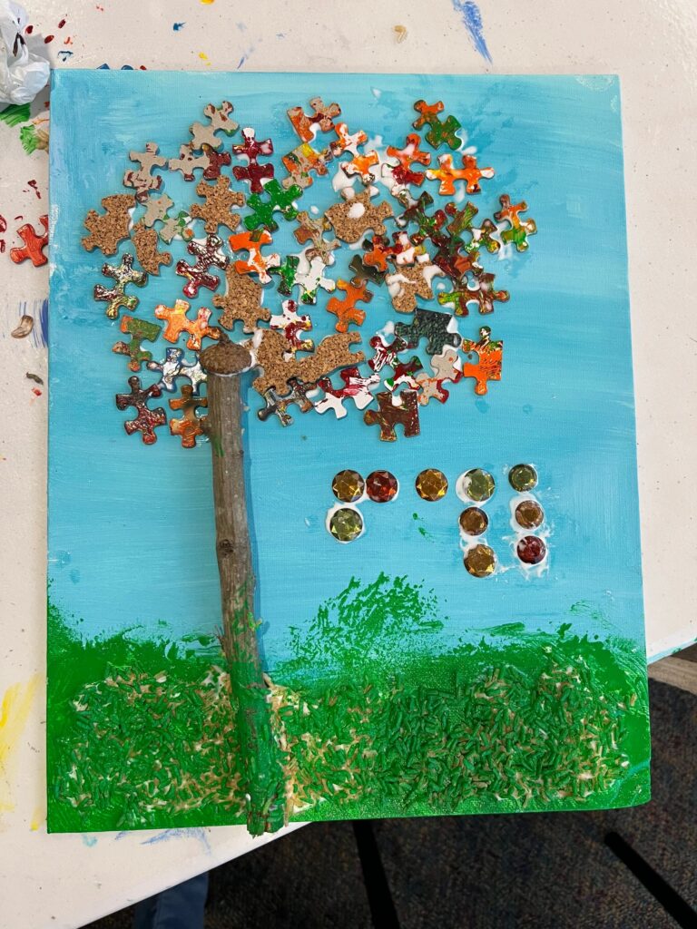 A fall tree canvas resting on the table, its canopy built from cork-backed puzzle pieces in gold, green and red, with amber gems clustered beside the twig trunk and green sprinkled grass below.