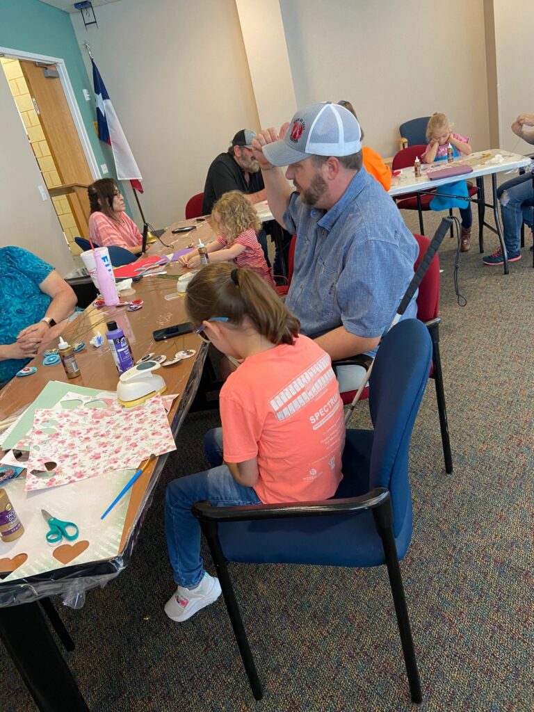 A volunteer helps an artist glue paper petals onto a wire stem at a table set with floral paper, scissors and paint.