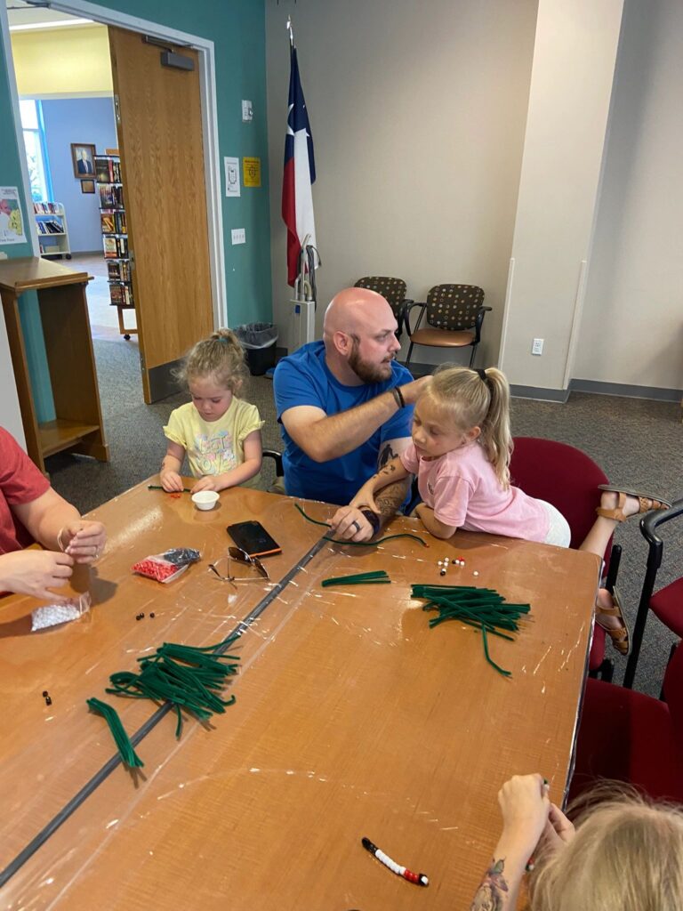 A volunteer helps two young artists at a table covered in green pipe cleaners and bowls of beads for a craft.