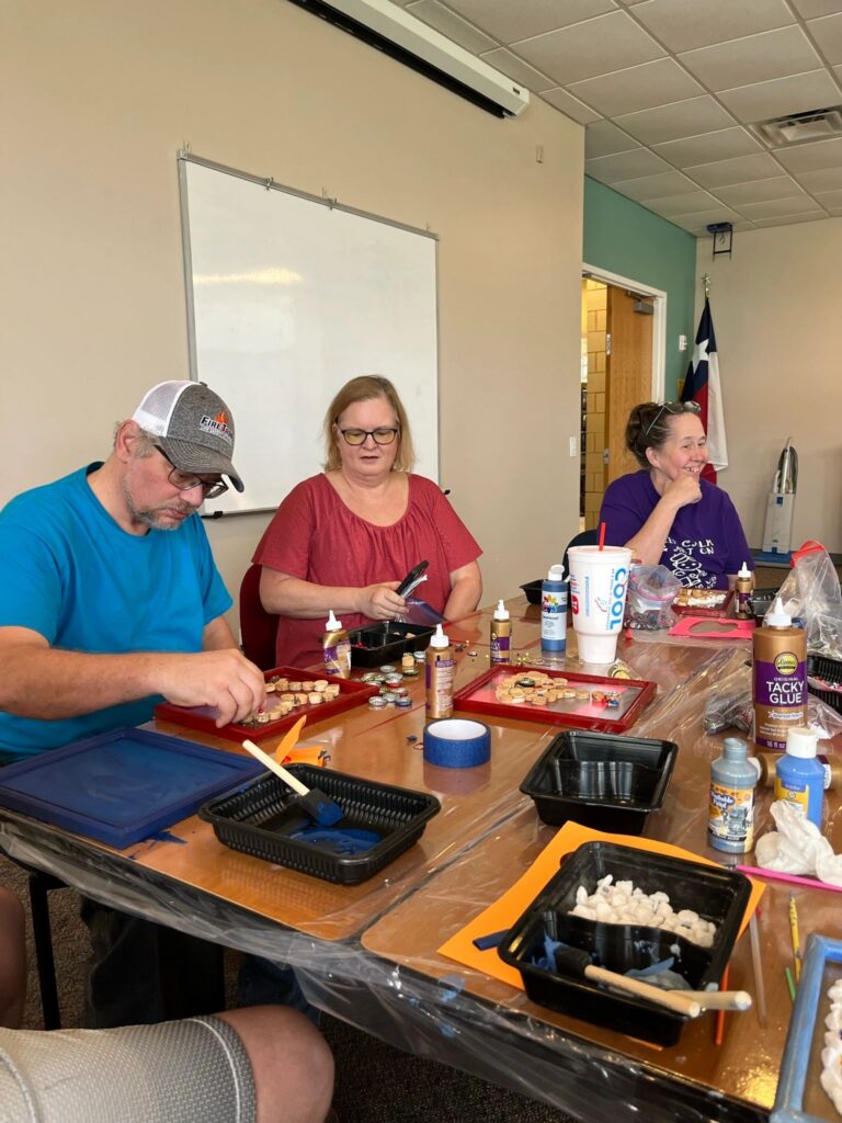 Artists and volunteers seated around a table covered in trays of corks and beads, gluing pieces onto framed boards.