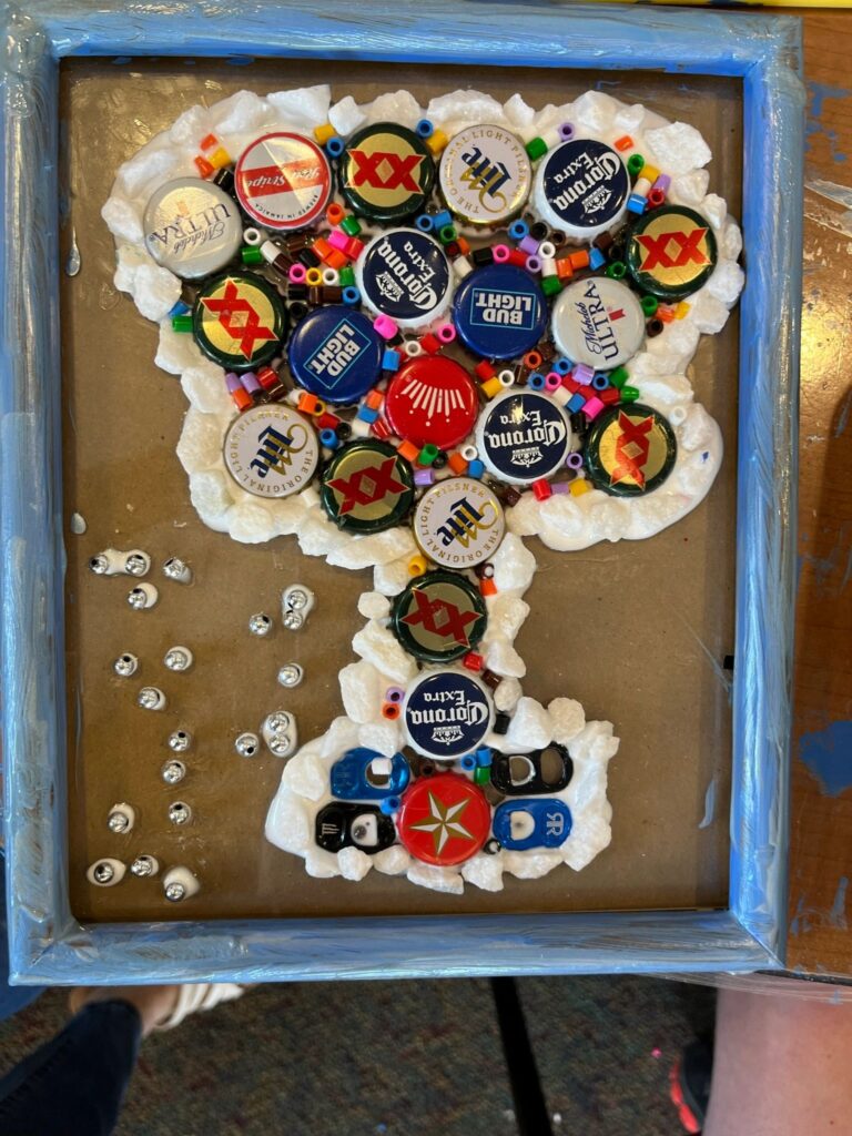 A finished cork art piece: a large number formed from corks and beer bottle caps arranged inside a blue frame, with loose silver beads scattered nearby.