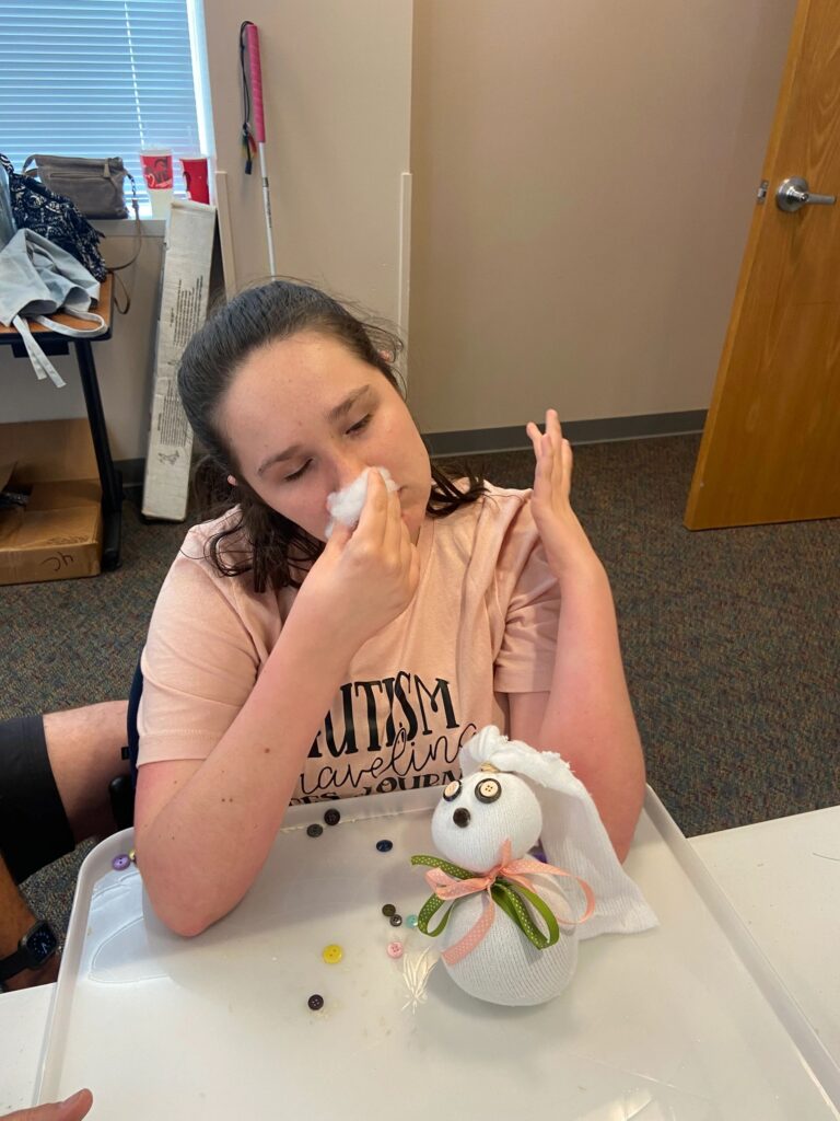 An artist wipes their face with a tissue at a table, a partly finished sock bunny decorated with a pink ribbon bow resting in front of them.