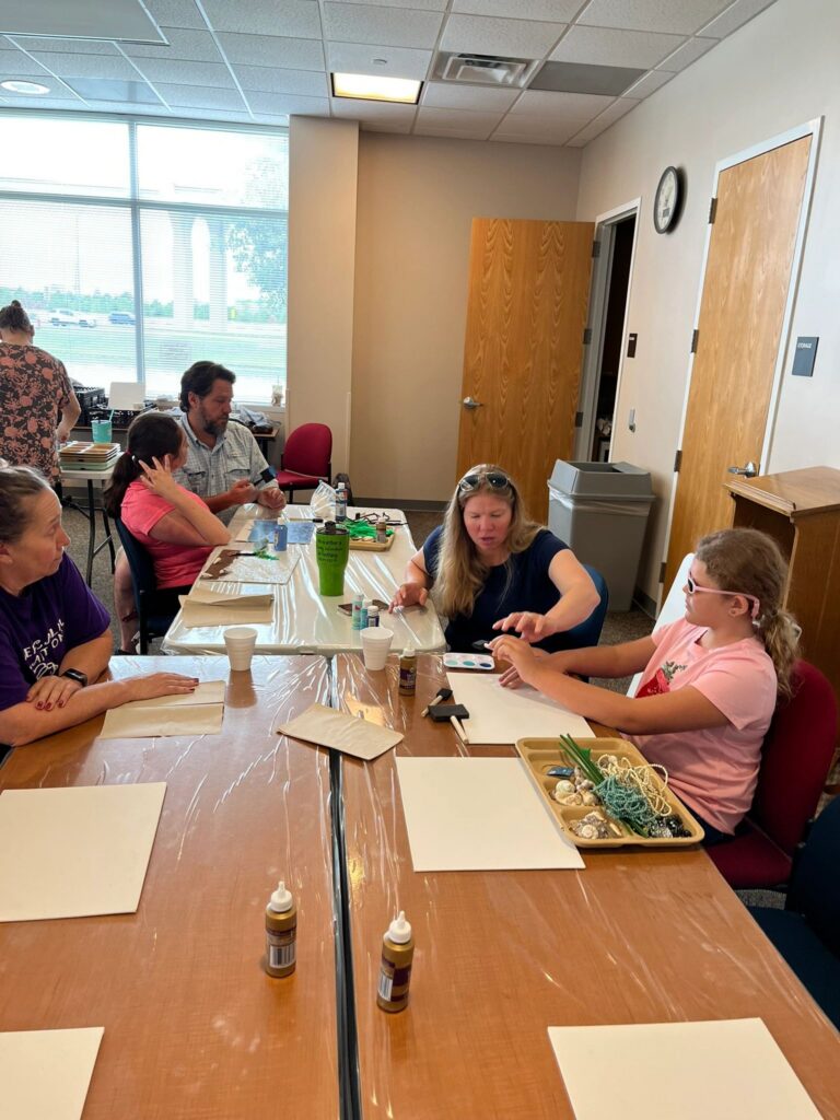A wide view of the studio, with artists and volunteers seated at tables working on blank and painted canvases, with craft supplies in trays.