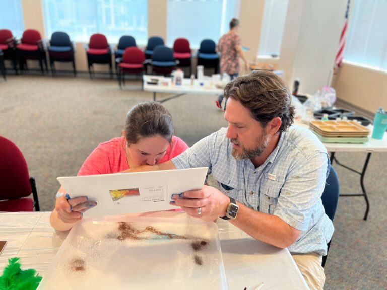 A volunteer helps an artist look closely at a finished canvas with a tray of sand nearby, in a room with rows of chairs in the background.