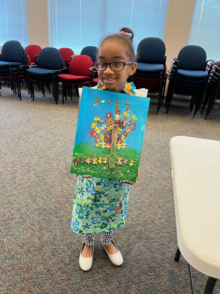 An artist holds up a fall tree canvas with a puzzle-piece canopy in red, yellow and gold, a real twig trunk, gold sequins scattered above and cork circles along the grass line.