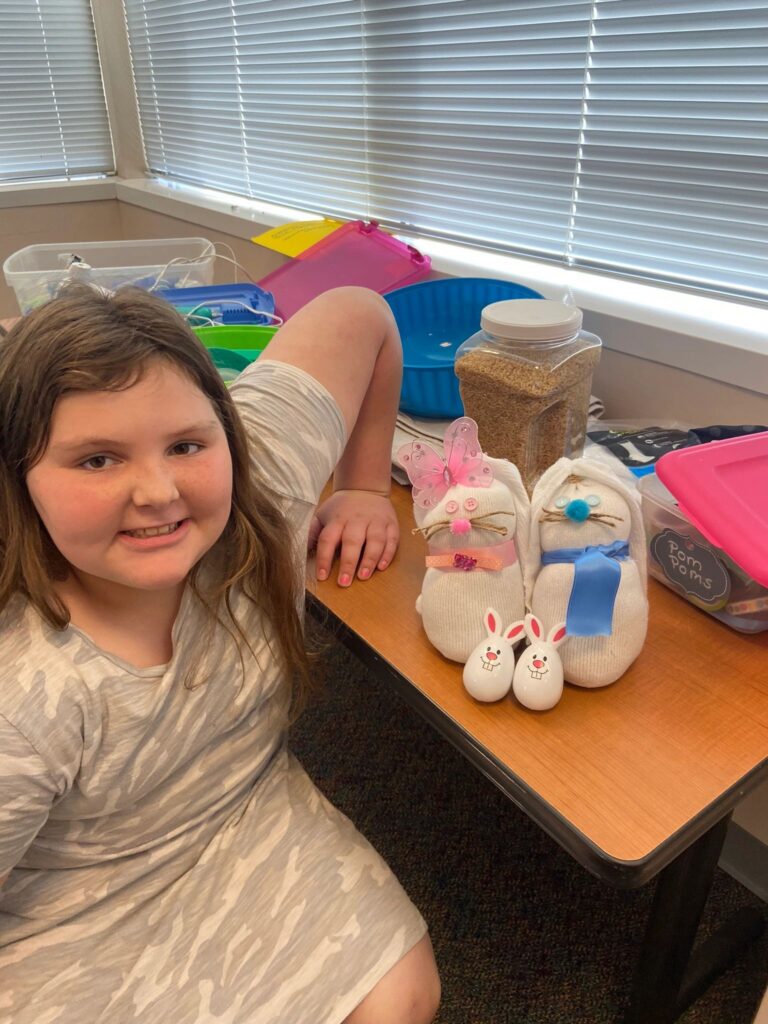 A finished pair of sock bunnies on a table, one with a pink butterfly and ribbon collar, the other with a blue bow, alongside two small bunny figurines.