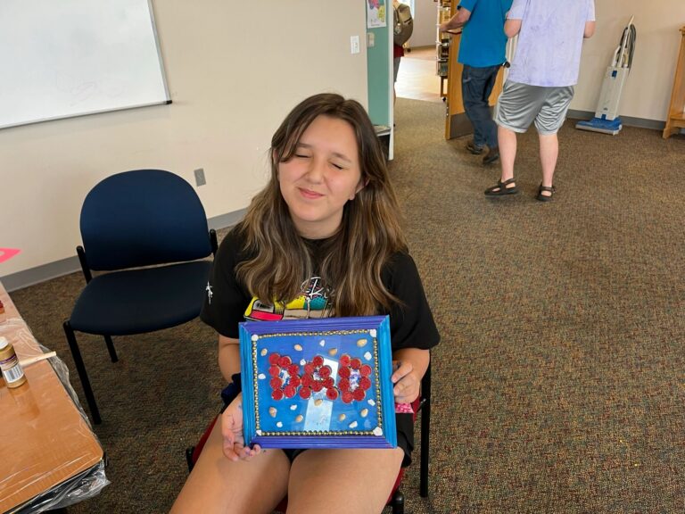 An artist holds up a framed piece spelling DAD in red corks on a blue background, decorated with small white and gold beads around the border.