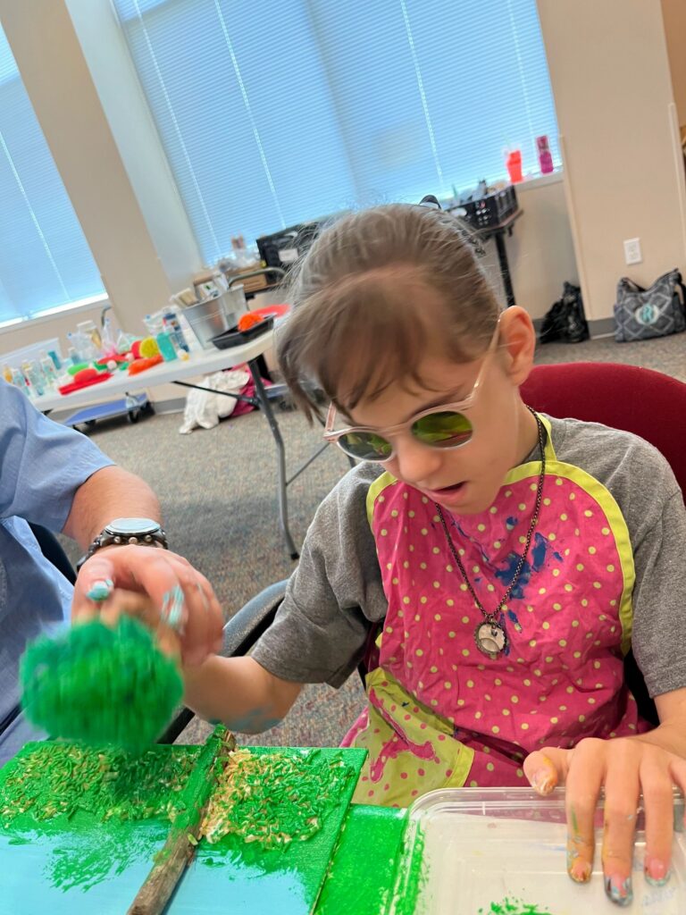 A volunteer helps an artist press a green textured pom-pom onto a canvas dotted with gold paint, with craft supplies spread on tables in the background.