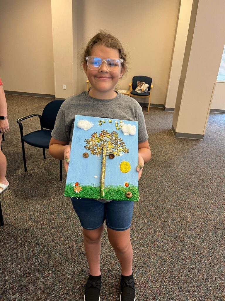 An artist holds up a fall tree canvas with a gold glitter canopy made from puzzle pieces, a twig trunk, cotton-wool clouds and a bright yellow glitter sun.