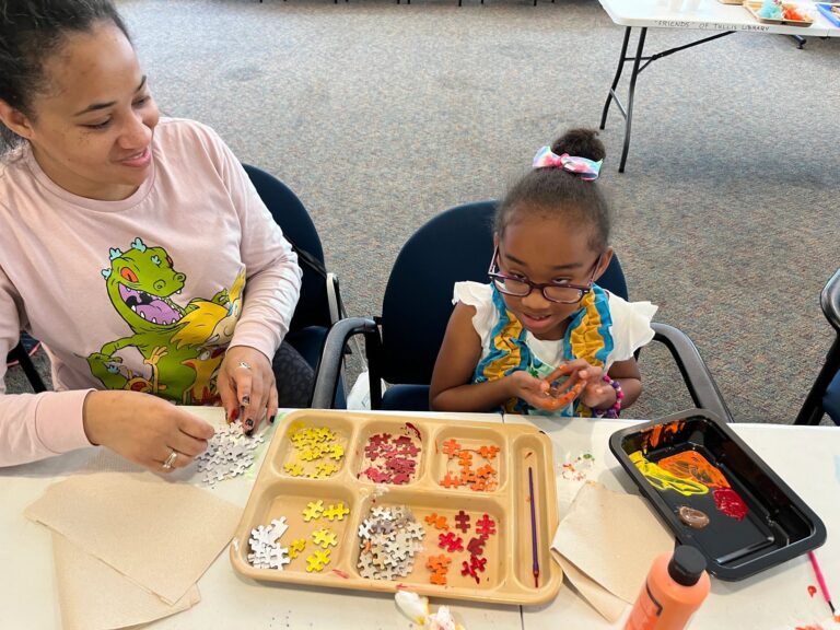An artist and a volunteer sort puzzle pieces into a divided tray by colour, working together at the table.
