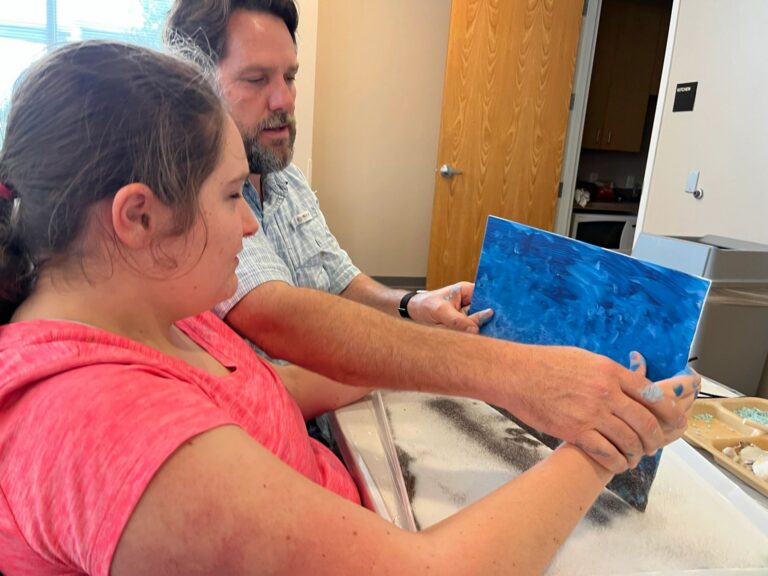 A volunteer helps an artist hold up a finished dark blue canvas, with a tray of shells and beads on the table.