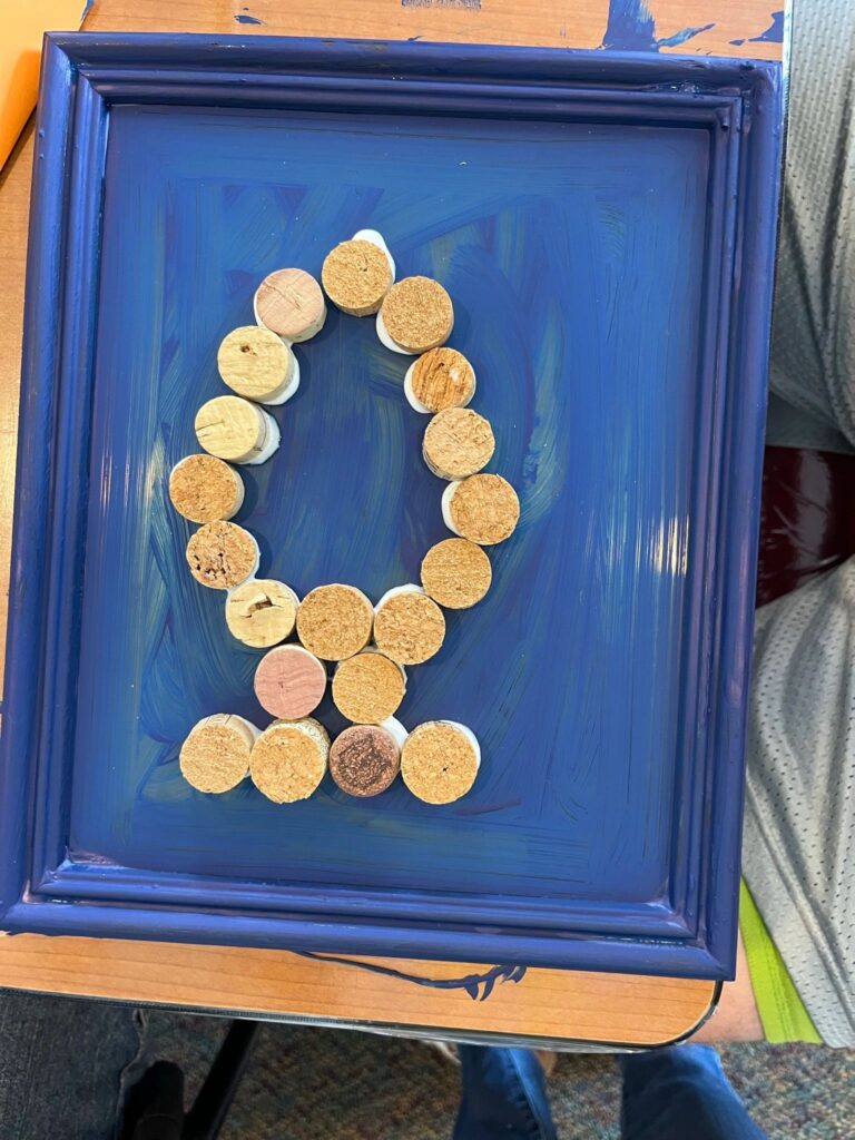 A framed cork piece in a blue frame showing the letter Q formed from wine corks standing upright against a painted blue background.