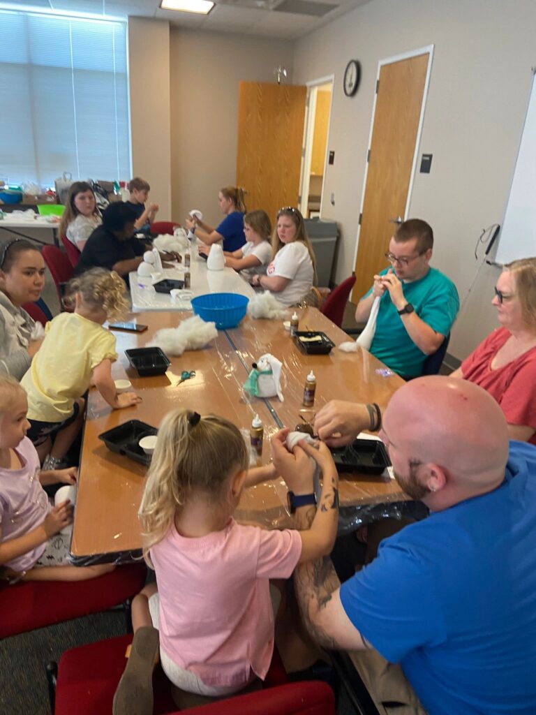 A wide view of the studio, with artists and volunteers seated at a long table making sock bunnies, cotton and craft supplies spread across the tabletop.