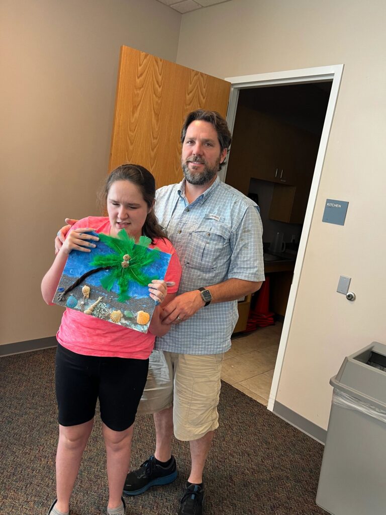 A volunteer and an artist hold up a finished canvas together, decorated with a feather palm tree, a twig trunk, seashells and sand along the base.