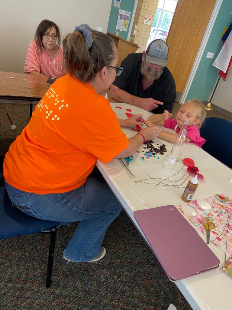 Two volunteers each hold a finished sock bunny wearing wire dragonfly antennae, one pink and one blue, standing near a table of craft supplies.
