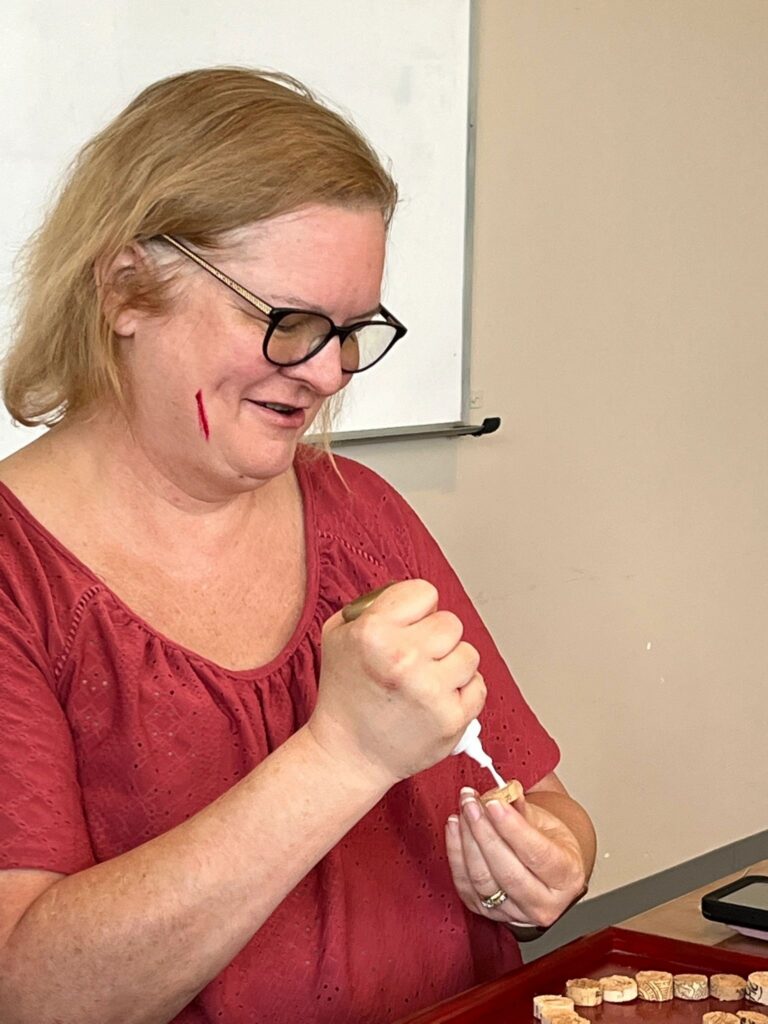 A volunteer glues a cork onto a small wooden piece at the table, with a tray of loose corks nearby.