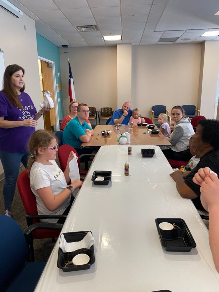 A volunteer speaks to a group of artists and volunteers gathered around a long table with sock bunnies, rice and glue supplies.