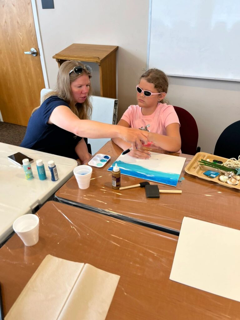 A volunteer helps an artist paint a row of blue dots above a dark blue painted band on a white canvas, with paint trays and a beaded tray nearby.