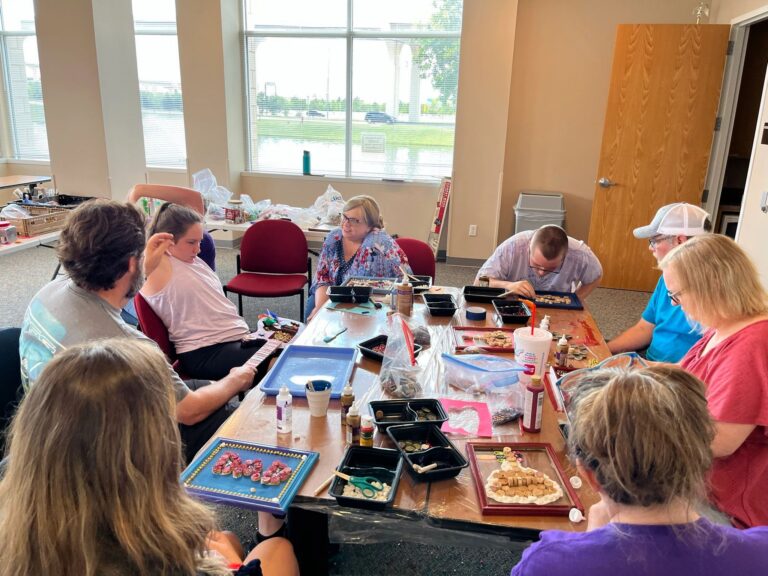 A wide view of the studio, with artists and volunteers seated around a table covered in trays of corks, beads and craft supplies.