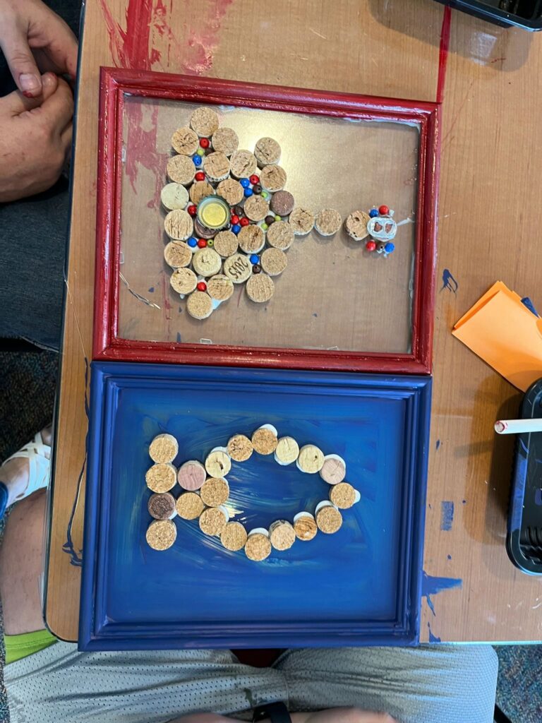 Two framed cork pieces side by side: the top in a red frame shows a cork airplane with beaded accents and the number 2025 spelled in cork, the bottom in a blue frame shows a fish shape made from corks on painted blue.