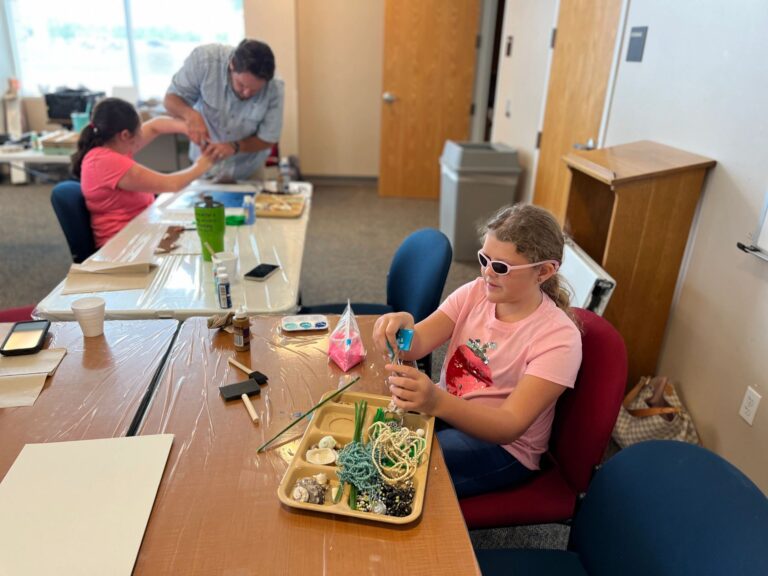A volunteer works with an artist at a table set with beaded strands, shells and craft supplies in trays.