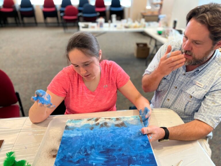 A volunteer helps an artist spread blue paint over their hands, with a textured blue and brown canvas on the table.